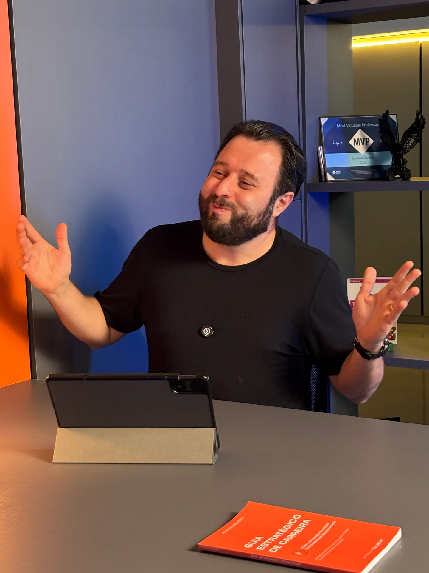 Joviano conversando à mesa, com a placa Microsoft MVP visível na estante ao fundo.