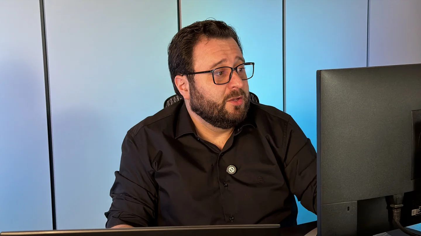 Joviano de óculos, dando aula diante dos monitores no estúdio.